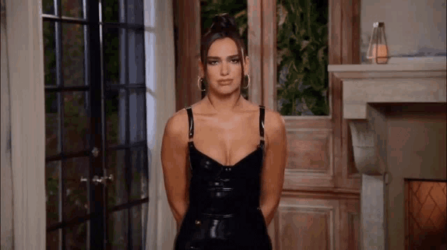 a woman in a black latex dress is standing in a room in front of a fireplace .