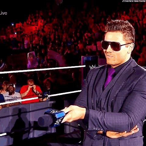 a man wearing sunglasses and a suit holds a microphone in front of a crowd .