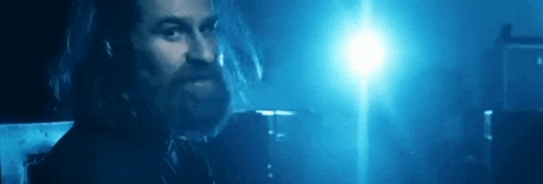 a man with long hair and a beard is standing in a dark room in front of a blue light .
