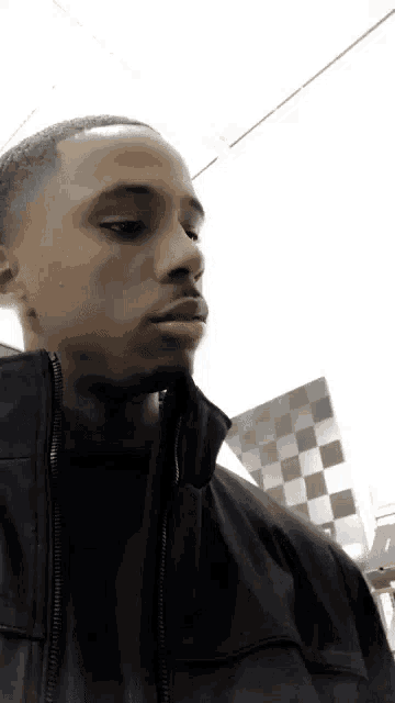 a man in a black jacket is standing in front of a checkered wall and looking at the camera .