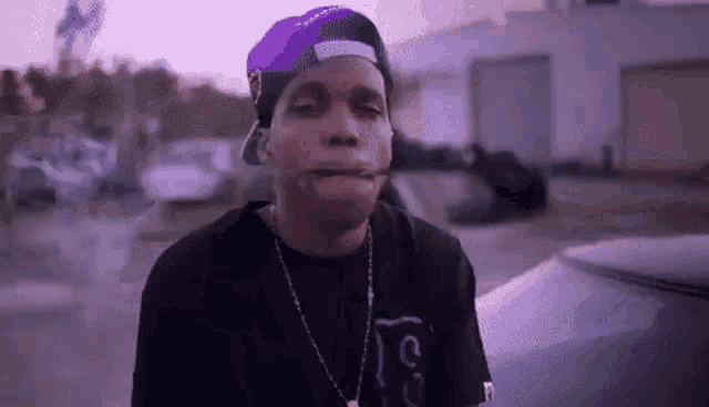 a man wearing a purple hat and a necklace is smoking a cigarette in front of a car .