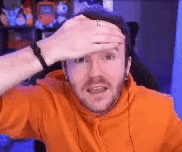 a man in an orange hoodie is holding his hand to his forehead and making a funny face .