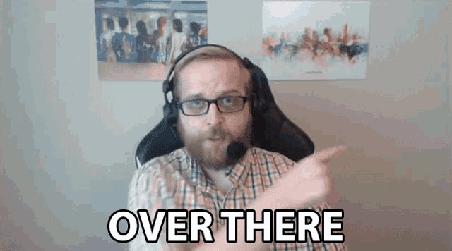 a man with glasses and headphones is pointing at the camera with the words over there behind him