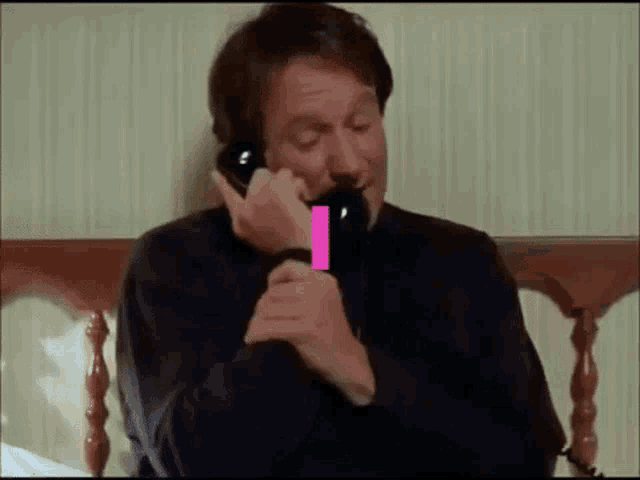 a man is sitting on a bed talking on a telephone with a pink ribbon coming out of his mouth .