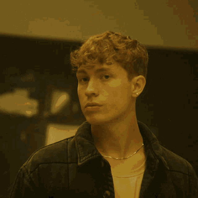 a young man with curly hair is wearing a denim jacket and a chain around his neck