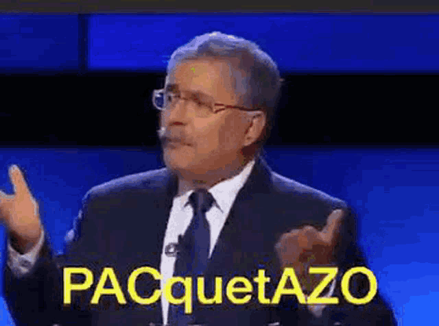 a man in a suit and tie is giving a speech on a television show while another man looks on .