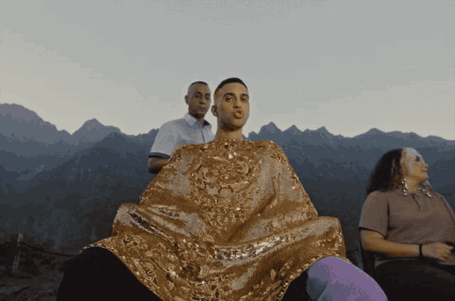 a man wearing a gold sequined dress stands in front of a mountain