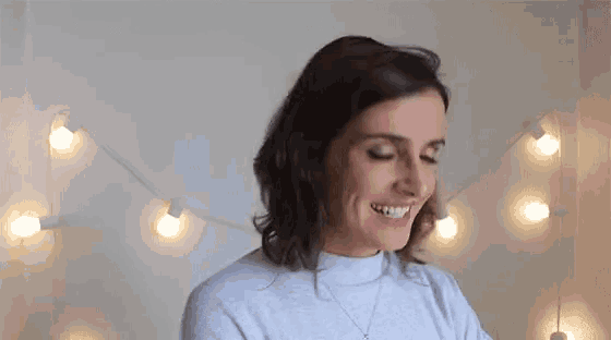 a woman wearing a grey sweater is smiling in front of a string of lights