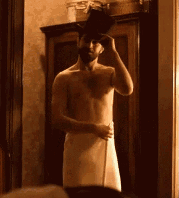 a shirtless man wearing a top hat and a towel is standing in front of a mirror .