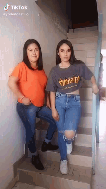 two women are standing next to each other on a set of stairs . one of them is wearing a thrasher shirt .