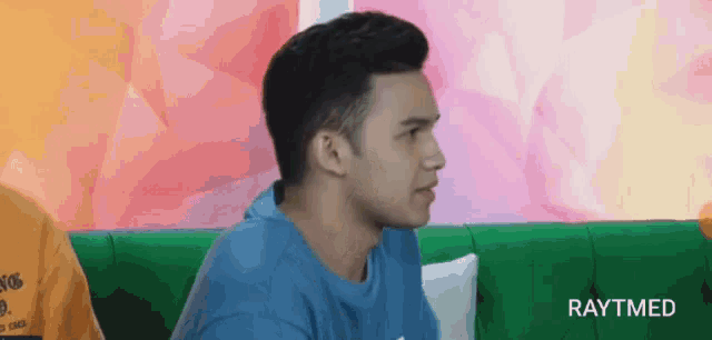 a man in a blue shirt is sitting on a green couch and looking at the camera .