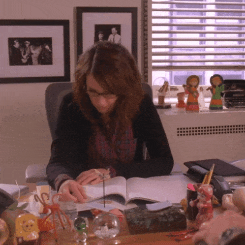 a woman sits at a desk reading a book surrounded by various objects