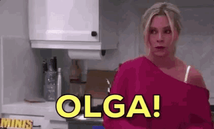 a woman in a pink sweater is standing in a kitchen with the word olga written in yellow letters .