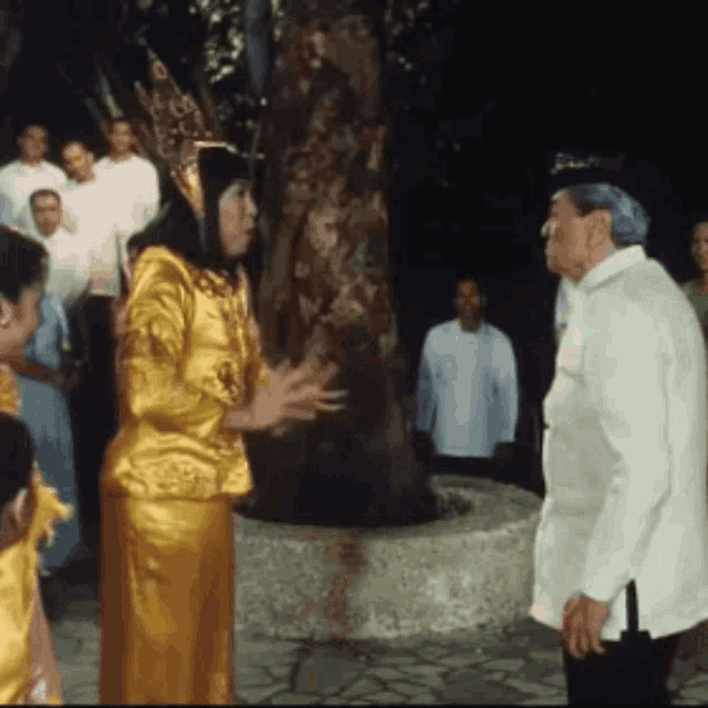 a woman in a gold dress talks to a man in a white shirt in front of a group of people