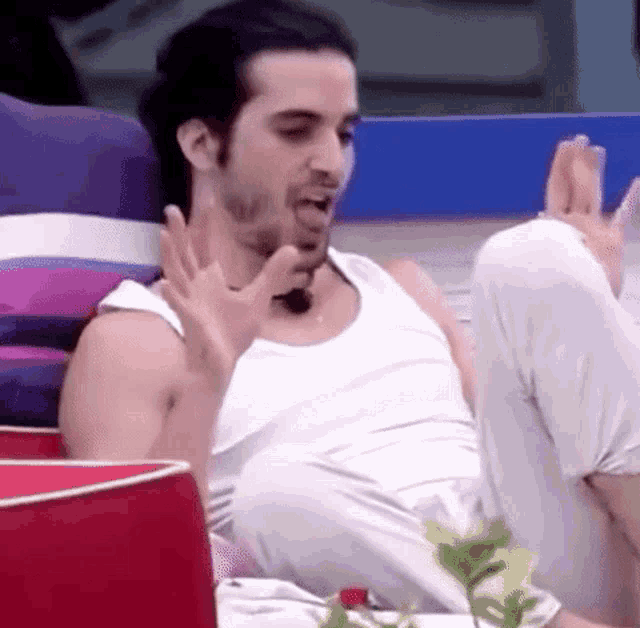a man in a white tank top is sitting on a couch with his feet up and making a funny face .