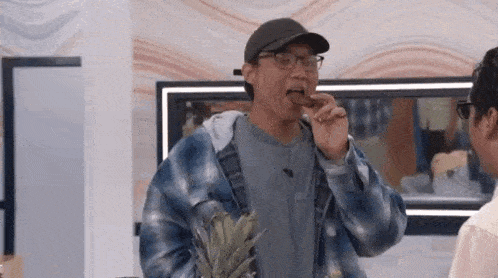 a man in a plaid shirt and hat is eating a pineapple while holding a pineapple .