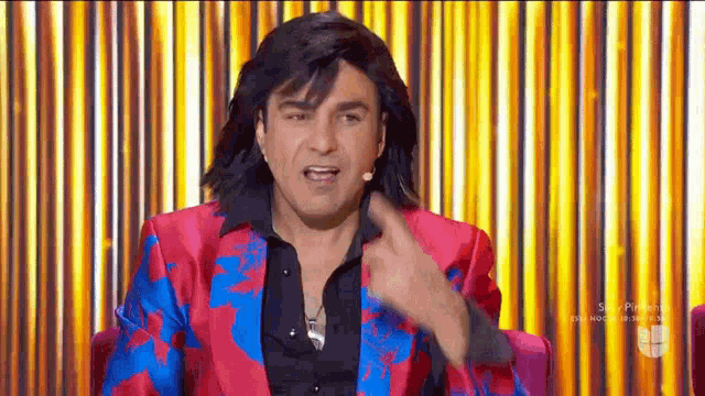 a man in a pink and blue suit is sitting in front of a yellow curtain and pointing at something .