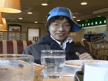 a man wearing a blue hat sits at a table in front of a green sign that says drink bar