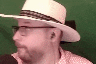 a man with a beard and glasses is wearing a cowboy hat and talking into a microphone .
