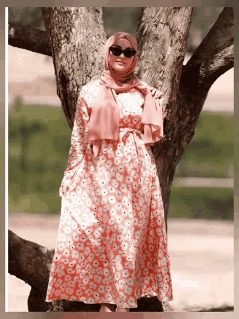 a woman wearing a pink floral dress and a pink scarf stands next to a tree