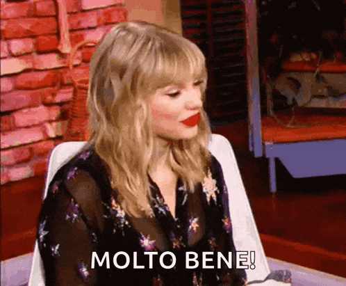 a woman with blonde hair and red lipstick is sitting in a chair and says molto bene !
