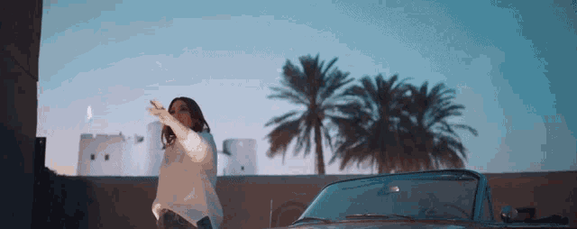 a woman in a white sweater is standing next to a car with palm trees in the background