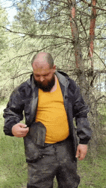 a bald man with a beard is standing in the woods holding a shovel