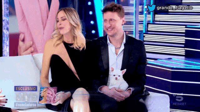 a man and a woman sit on a couch with a small white dog in their lap and the words esclusivo on the bottom right