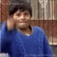 a young boy in a blue sweater is waving his hand in front of a fence .