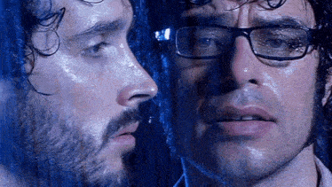 a man with glasses and a beard is looking at his reflection in a window