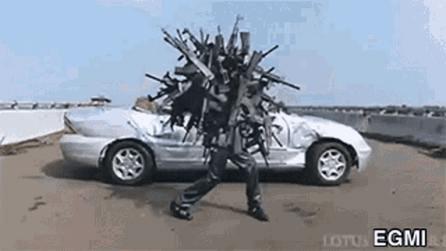 a man is carrying a bunch of guns on his head in front of a silver car