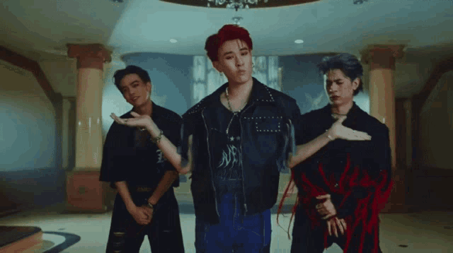 a group of young men are standing in a room and one of them is wearing a black jacket with studs
