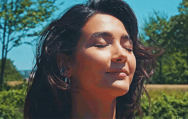 a close up of a woman 's face with her eyes closed in front of trees and a blue sky