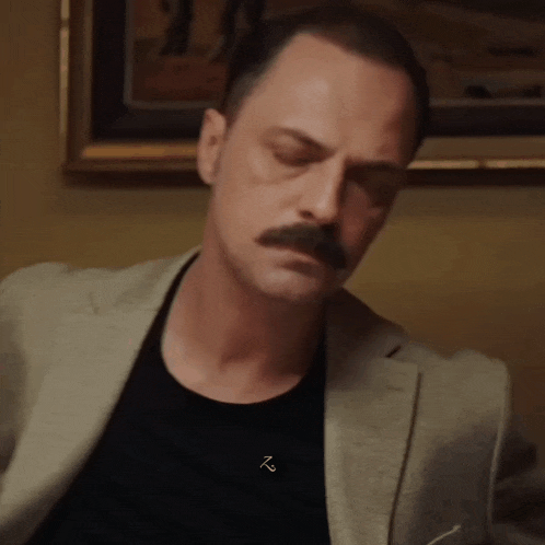 a man with a mustache is wearing a black t-shirt and a tan jacket .
