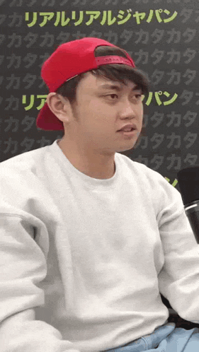 a man wearing a white sweater and a red hat is sitting in front of a wall with chinese characters on it