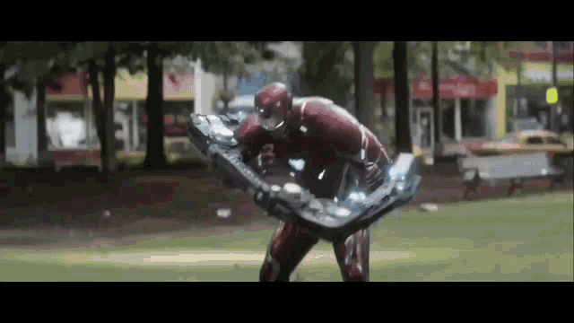 a man in a superhero costume is standing in a park holding a giant shield .