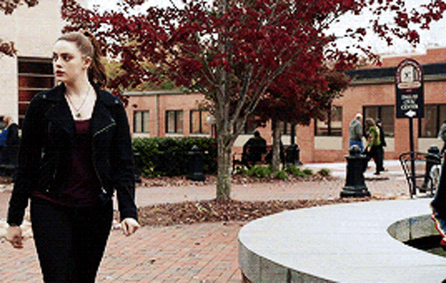 a woman in a black jacket is walking down a sidewalk in front of a building with a clock on it