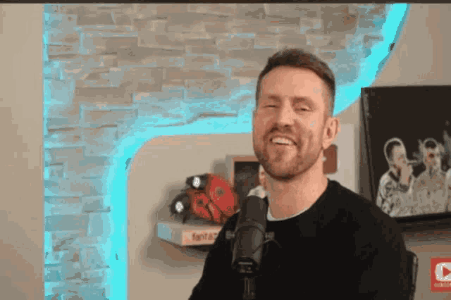 a man with a beard is smiling in front of a microphone in front of a brick wall