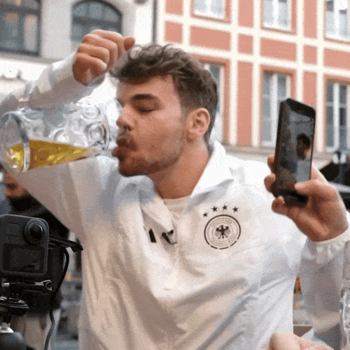 a man wearing a white shirt with a german emblem on it drinks from a pitcher