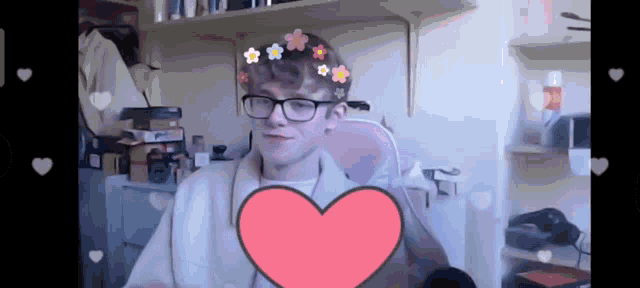a young man wearing glasses and a flower crown is holding a pink heart in front of him .