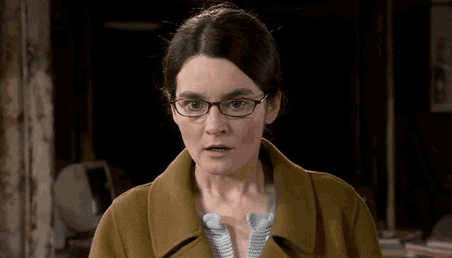 a woman wearing glasses and a brown coat is looking at the camera
