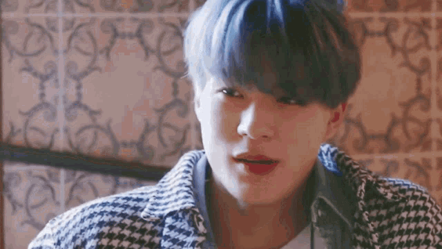a close up of a young man with blue hair wearing a plaid jacket and looking at the camera .