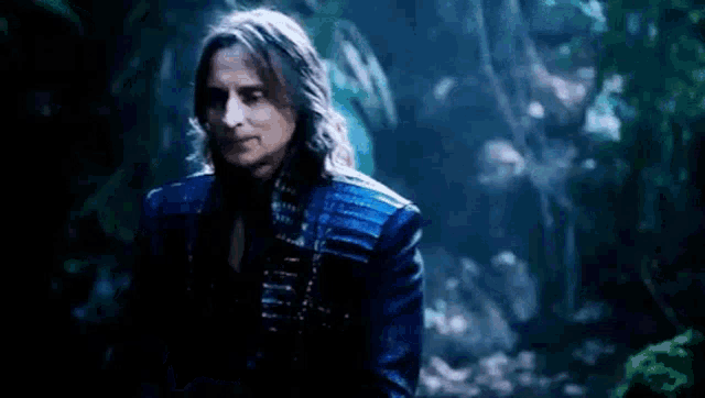 a man with long hair is wearing a blue jacket and standing in the woods .