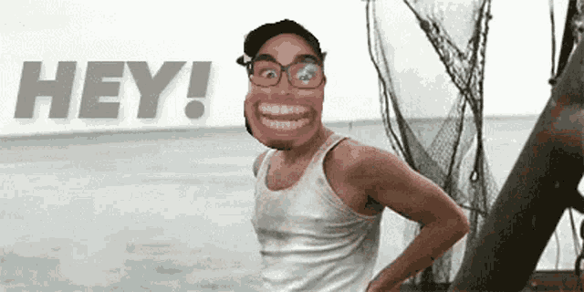 a man with glasses and a pink face is standing in front of the ocean and says hey