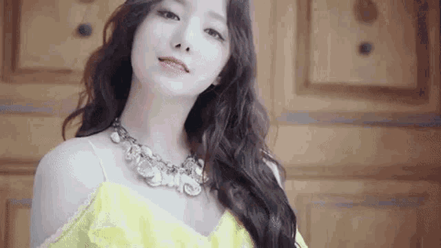 a woman in a yellow dress and necklace is standing in front of a wooden wall .