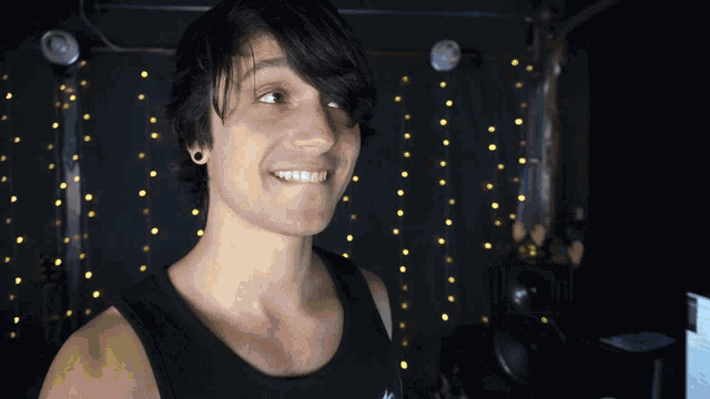 a man in a black tank top smiles in front of string lights
