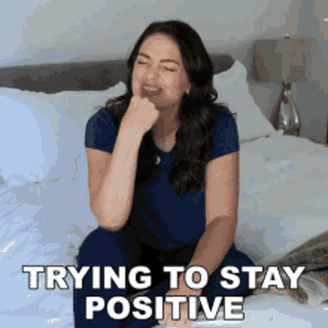 a woman is sitting on a bed with the words " trying to stay positive " on the bottom