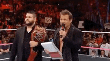 two men are standing in a wrestling ring talking into microphones . one of the men is holding a championship belt .