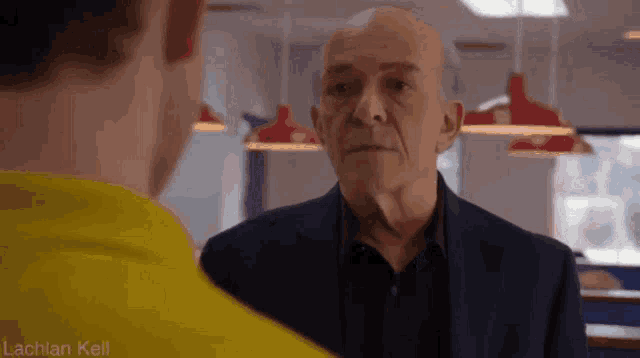 an older man in a suit talks to a younger man in a yellow shirt in a diner .