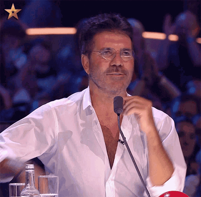 a man wearing glasses and a white shirt is speaking into a microphone in front of a crowd